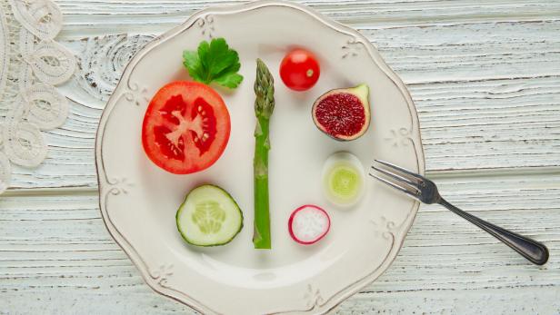 Verschiedene Gemüsescheiben, eine Feige und ein Spargel liegen dekorativ auf einem weißen Teller mit einer kleinen Gabel.