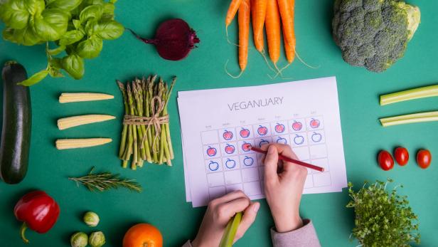 Verschiedene Gemüsesorten liegen um einen Kalender für Veganuary, in den eine Person mit einem Stift einträgt.