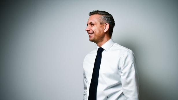 Ein Mann im weißen Hemd und dunkler Krawatte, Wirtschaftsminister Hattmannsdorfer, steht seitlich und lächelt vor einer hellgrauen Wand.