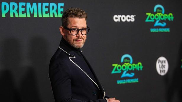 Ein Mann mit Brille und Bart steht vor einer Wand mit dem Zootopia-2-Logo und schaut zur Seite.