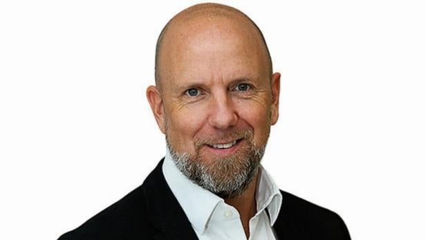 Ein Mann mit Glatze, grauem Bart und Anzug lächelt freundlich vor weißem Hintergrund.