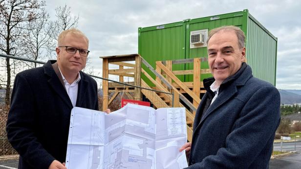 Zwei Männer im Mantel halten gemeinsam einen großen Bauplan vor einem grünen Container mit Holztreppe.