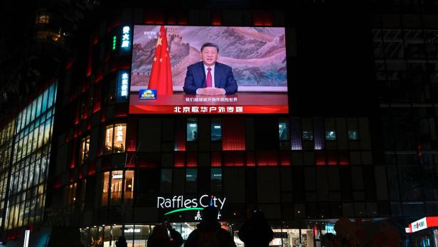 Menschen stehen vor einem Einkaufszentrum, über dessen Eingang eine große Leinwand mit einer Ansprache gezeigt wird.