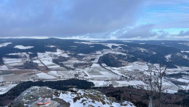 Verschneite Felder und Dörfer in einem weiten Tal, im Vordergrund Felsen mit etwas Schnee und kahlen Ästen.