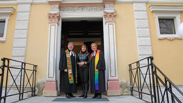 Drei Personen stehen vor dem Eingang einer Kirche, zwei davon tragen schwarze Talare mit bunten Stolen.