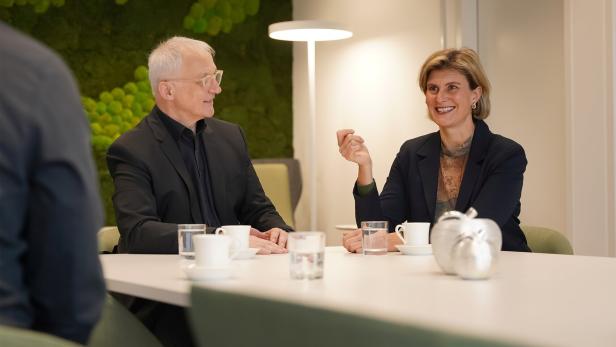 Zwei Personen im Gespräch an einem Tisch mit Tassen, Gläsern und silbernen Dekoräpfeln, beide wirken freundlich.