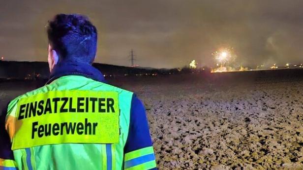 Eine Person in Feuerwehrkleidung mit der Aufschrift „Einsatzleiter Feuerwehr“ steht nachts auf einem Feld und blickt auf einen Brand in der Ferne.