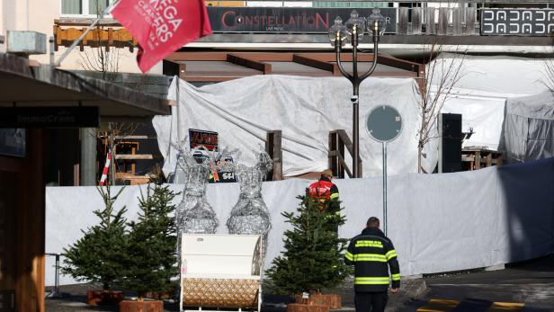 Zwei Personen in Warnwesten gehen an abgedeckten Bereichen vorbei, umgeben von Tannenbäumen und Drahtfiguren von Rentieren.