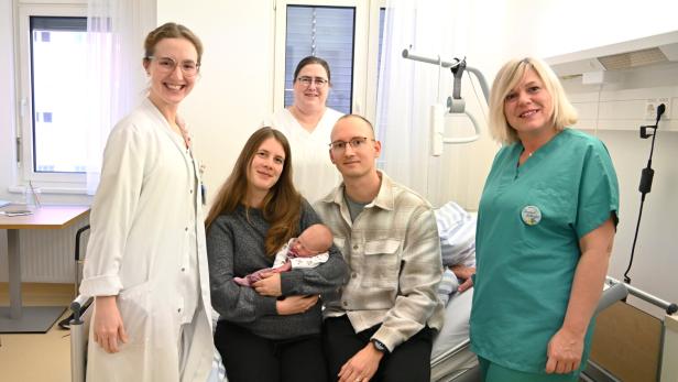 Eine Frau hält ein Neugeborenes im Arm, neben ihr sitzt ein Mann, umgeben von medizinischem Personal in einem Krankenhauszimmer.