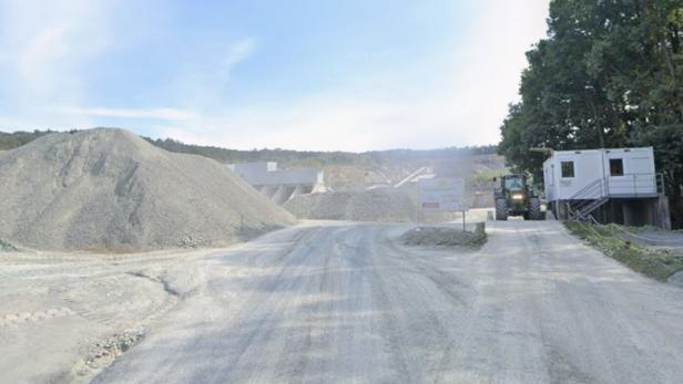 Große Schotterhaufen, ein Traktor und Container stehen auf einem staubigen Gelände neben Bäumen unter blauem Himmel.
