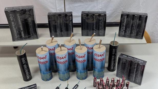 Mehrere Feuerwerkskörper verschiedener Größen auf einem Tisch vor einem Schild mit Ortsangabe.