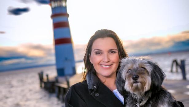 Barbara Karlich steht mit einem Hund im Arm vor einem Leuchtturm an einem See