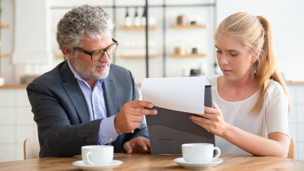 Eine junge Frau und ein Man sitzen an einem Tisch und sehen sich ein Dokument auf einem Klemmbrett an.