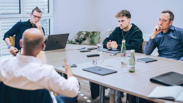 Vier Männer sitzen an einem Tisch und führen eine konzentrierte Besprechung mit Laptops, Getränken und Notizbüchern.