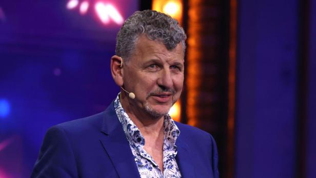 Ein Mann mit Mikrofon im blauen Anzug gestikuliert vor farbigem Hintergrund.