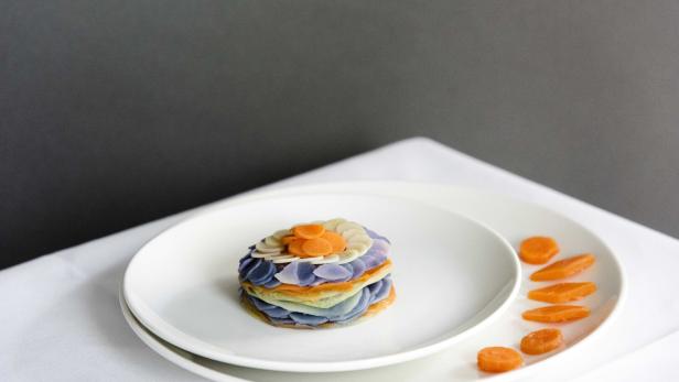 Auf einem Teller liegt ein kunstvoll geschichteter, farbenfroher Gemüseturm, dekoriert mit Karottenscheiben.