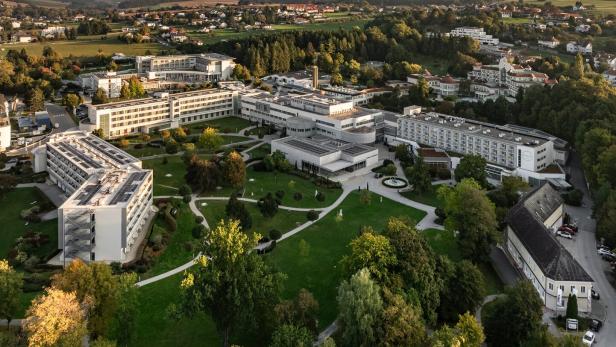 Mehrere moderne Gebäude mit weißen Fassaden liegen inmitten einer weitläufigen, grünen Parkanlage mit Wegen und Bäumen.