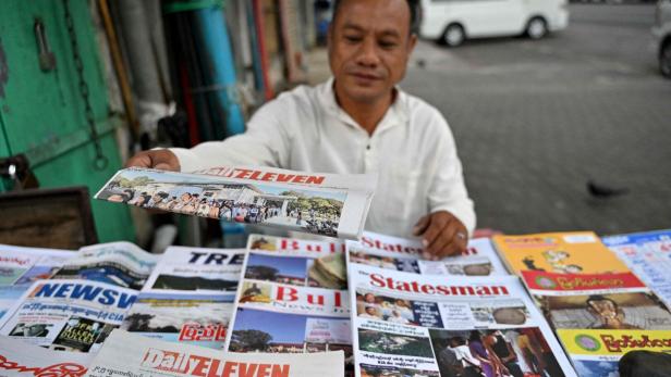 Ein Mann verkauft verschiedene Zeitungen und Magazine an einem Straßenstand und hält eine Zeitung in der Hand.