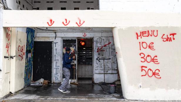 Ein Mann mit Besen steht vor einem mit Graffiti besprühten Hauseingang, an der Wand stehen Preise in roter Schrift.