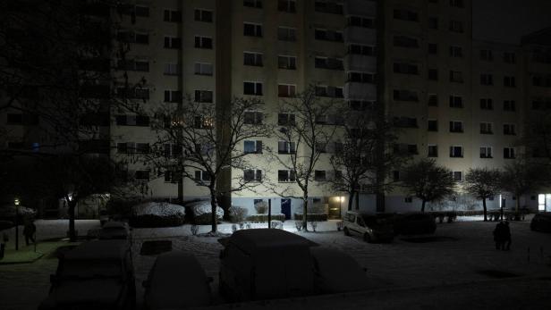 Vor einem mehrstöckigen Wohnhaus stehen schneebedeckte Autos und Bäume in einer dunklen, winterlichen Nacht.