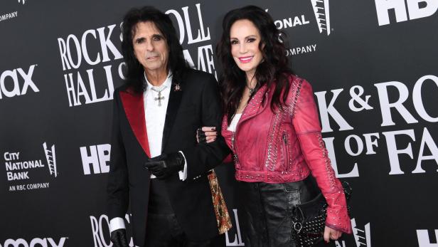 Ein Mann im schwarzen Anzug mit roten Details und eine Frau in pinker Lederjacke posieren gemeinsam vor einer Wand mit Schriftzug.