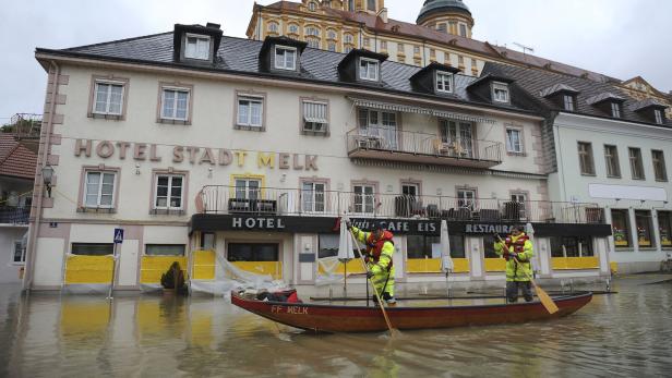 Zwei Einsatzkräfte fahren mit einem Boot durch eine überflutete Straße vor einem Hotel, im Hintergrund ein barockes Gebäude.