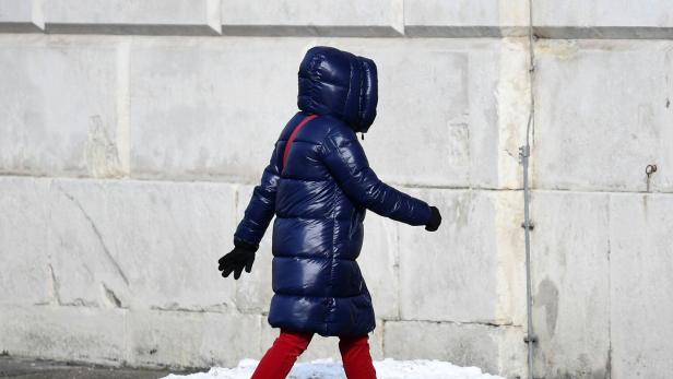 Person mit langer, dicker, blauer Daunenjacke, roter Hose und grauen Schuhen geht an einer schneebedeckten Straße entlang.