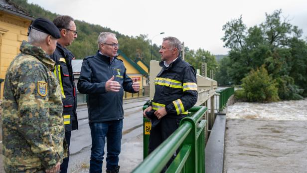 Vier Männer in Uniformen stehen auf einer Brücke und besprechen die Hochwassersituation am reißenden Fluss.