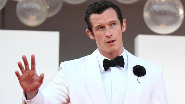 Ein Mann im weißen Smoking mit schwarzer Fliege hebt die Hand und steht vor einer festlichen Kulisse.