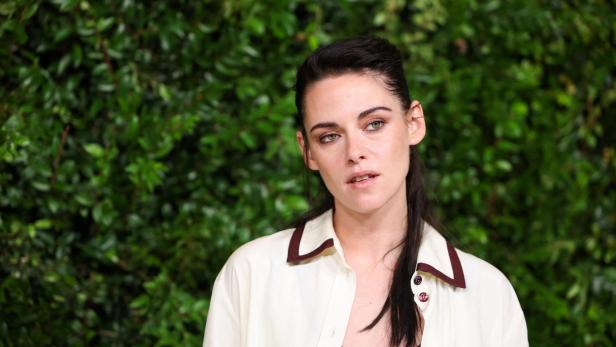 Kristen Stewart trägt ein cremefarbenes Hemd mit braunen Akzenten vor einer grünen Hecke.