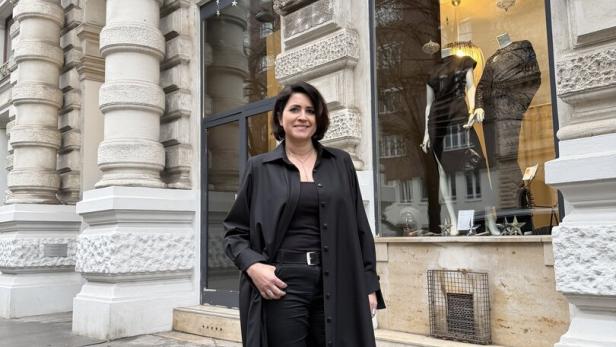 Eine Frau in schwarzer Kleidung steht vor einem Modegeschäft mit Schaufensterpuppen und orangefarbenem Schild auf nasser Straße.