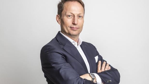 Mann im dunklen Anzug mit verschränkten Armen steht vor hellem Hintergrund und blickt selbstbewusst nach vorn.