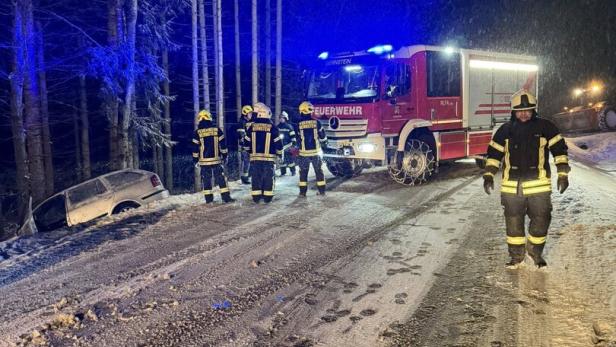 Feuerwehrleute sichern bei Nacht eine verschneite Straße, während ein Auto im Straßengraben liegt.
