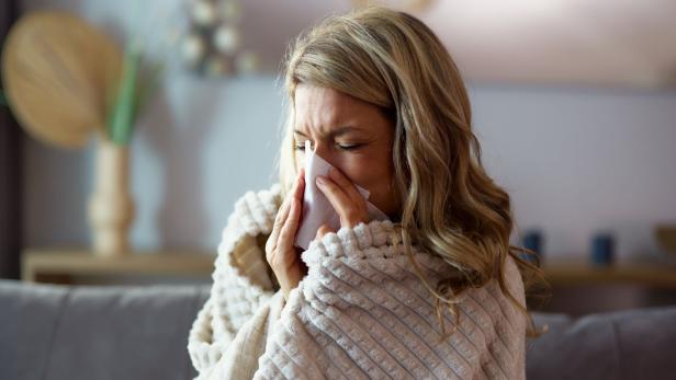 Ill woman covered with blanket blowing her nose at home.