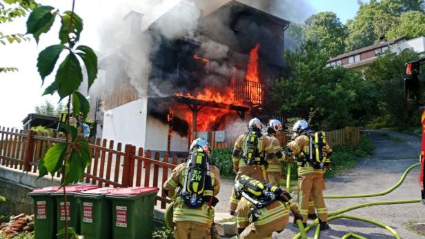 Feuerwehrleute bekämpfen einen Hausbrand, Flammen schlagen aus dem Gebäude und Rauch steigt auf.