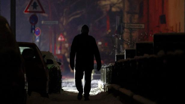 Eine Person geht nachts auf einem verschneiten Gehweg zwischen parkenden Autos und Häusern.