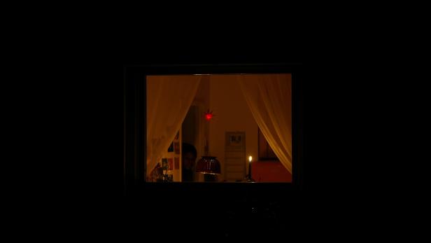 Ein Fenster mit Vorhängen zeigt einen schwach beleuchteten Raum, in dem eine Kerze leuchtet.
