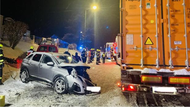 Ein silbernes Auto ist frontal mit einem orangefarbenen Lkw kollidiert, Feuerwehrleute und Einsatzfahrzeuge sind vor Ort.