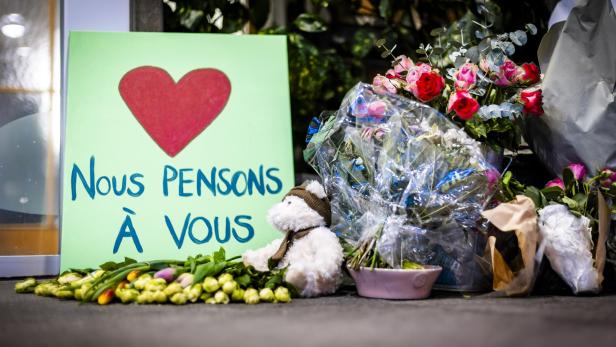 Ein Schild mit Herz und "Nous pensons à vous" liegt neben Blumen und einem Teddybär.