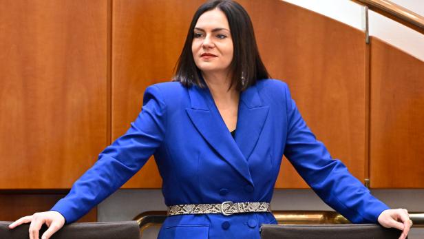 Frau mit schulterlangem dunklem Haar trägt einen blauen Blazer und steht mit ausgebreiteten Armen an zwei Stühlen.