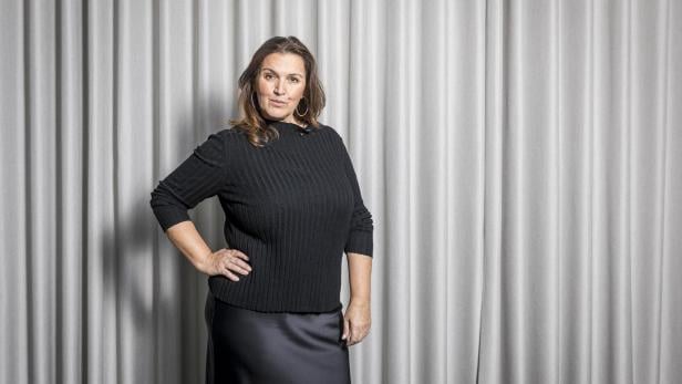 Frau mit langen braunen Haaren trägt schwarzen Pullover und Rock, steht vor einem hellgrauen Vorhang.