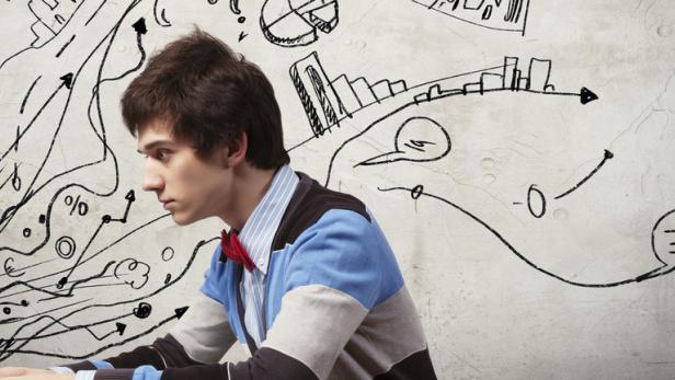 Ein junger Mann mit Fliege arbeitet konzentriert am Laptop vor einer Wand mit Skizzen.