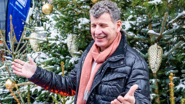 Ein Mann mit schwarzer Jacke und rotem Schal steht lachend zwischen geschmückten, verschneiten Tannenbäumen.