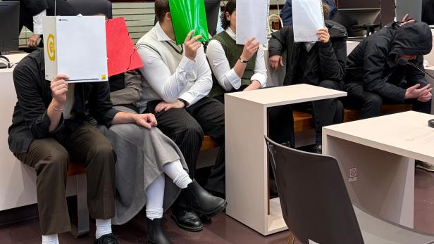 Mehrere Personen verdecken ihre Gesichter mit Aktenordnern und Papier in einem Gerichtssaal.