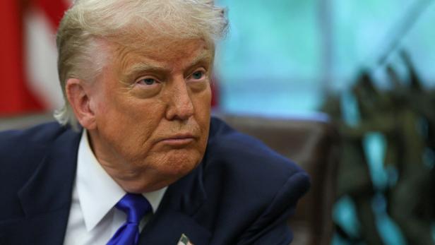 Donald Trump blickt mit ernstem Gesichtsausdruck zur Seite, im Hintergrund eine US-Flagge.