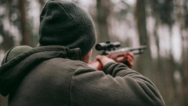 Eine Person mit Mütze und Jacke zielt mit einem Gewehr im Wald.