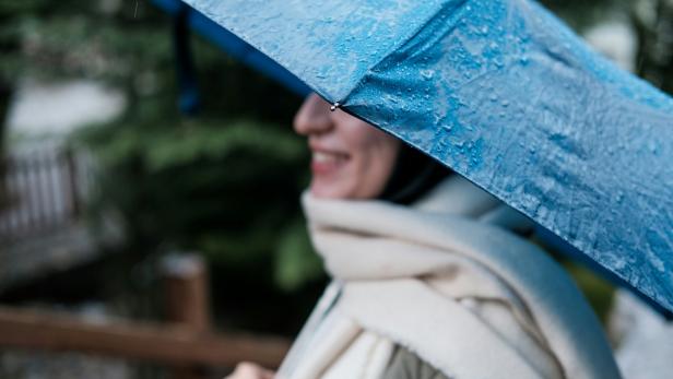 Person mit Schal und blauem Regenschirm im Regen, unscharfer Hintergrund mit Grün.