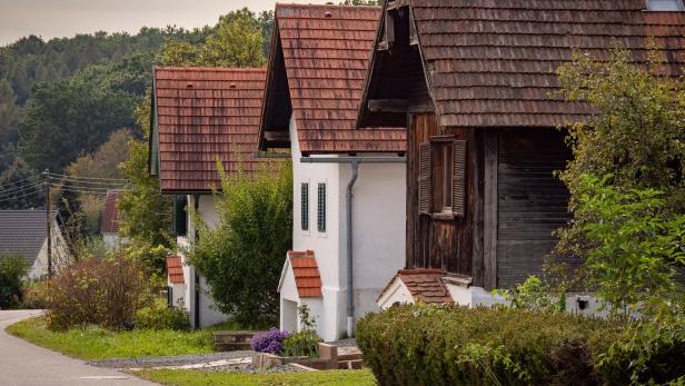 Mehrere kleine Häuser mit roten Ziegeldächern stehen nebeneinander an einer ruhigen Straße, umgeben von viel Grün.