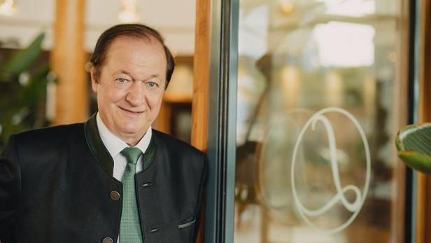 Ein älterer Mann im Trachtenanzug mit grünem Schlips steht lächelnd neben einer Glastür mit verziertem Logo.