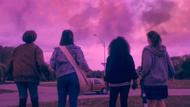 Vier Jugendliche stehen nebeneinander und blicken unter einem pink-violetten Himmel auf eine Straße mit geparktem Auto.